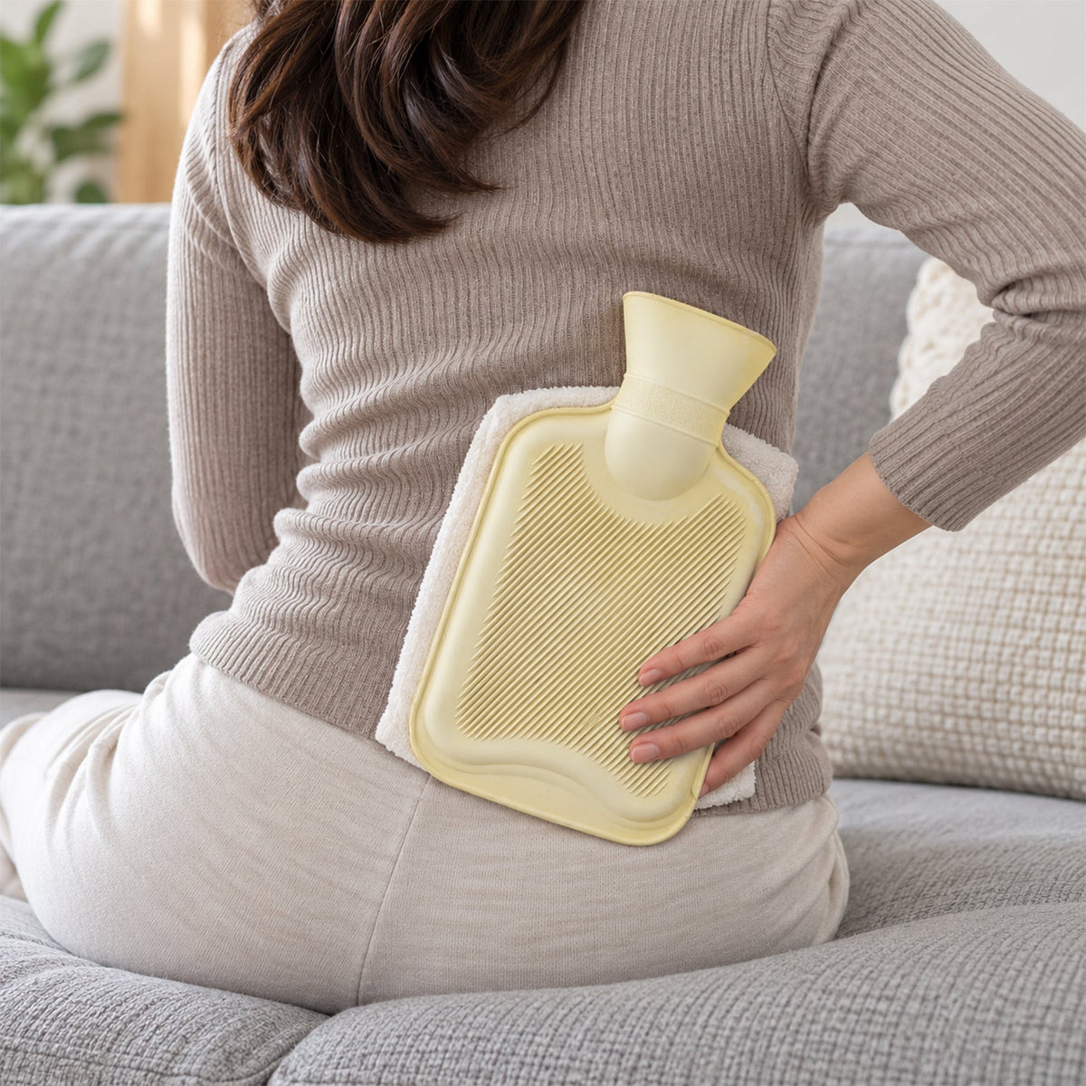 A person sits on a gray sofa, holding a light yellow hot water bottle wrapped in a white towel against their lower back.