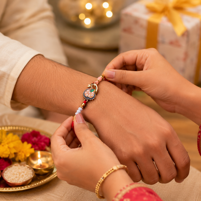 Hands tie a red thread with a gold peacock charm onto a man's wrist, with a gift and flowers in the background.
