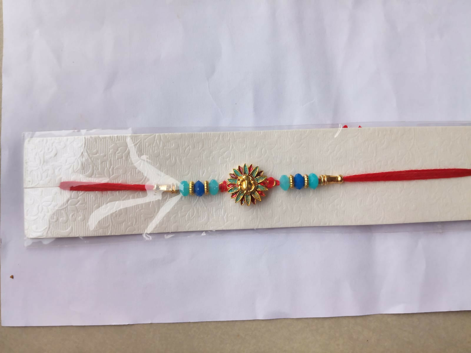 A red thread bracelet with a gold Ganesh charm, blue and light blue beads, and gold accents, displayed on a white textured
