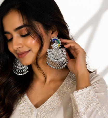 A woman with dark hair wears a large silver peacock earring with colorful details and sparkling gems.