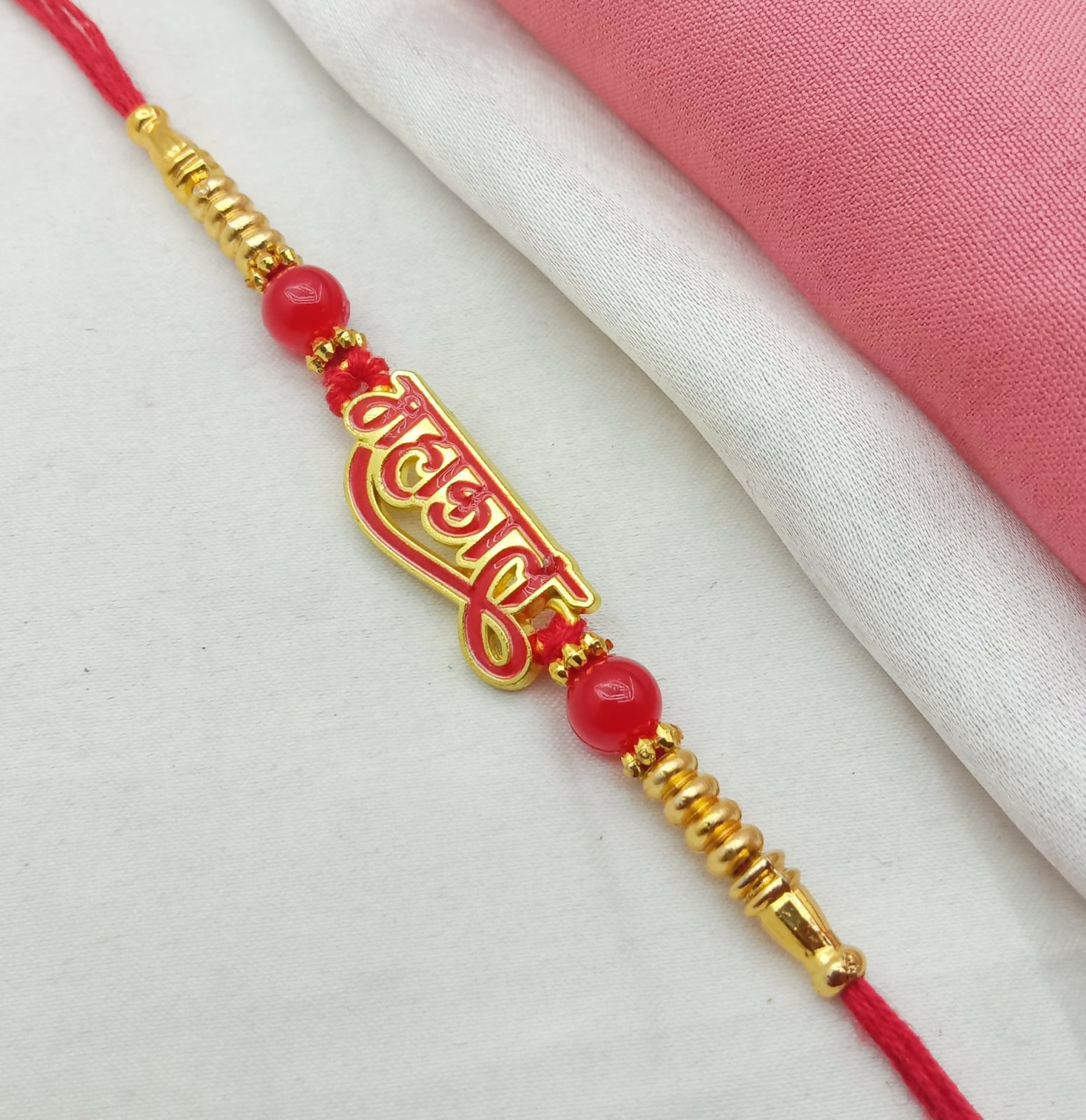 A red thread with a gold and red Mahakal calligraphy charm, red beads, and gold accents lies on a white and pink fabric