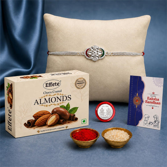 A silver-colored Rakhi with a central floral design featuring green and red accents rests on a beige cushion, surrounded by