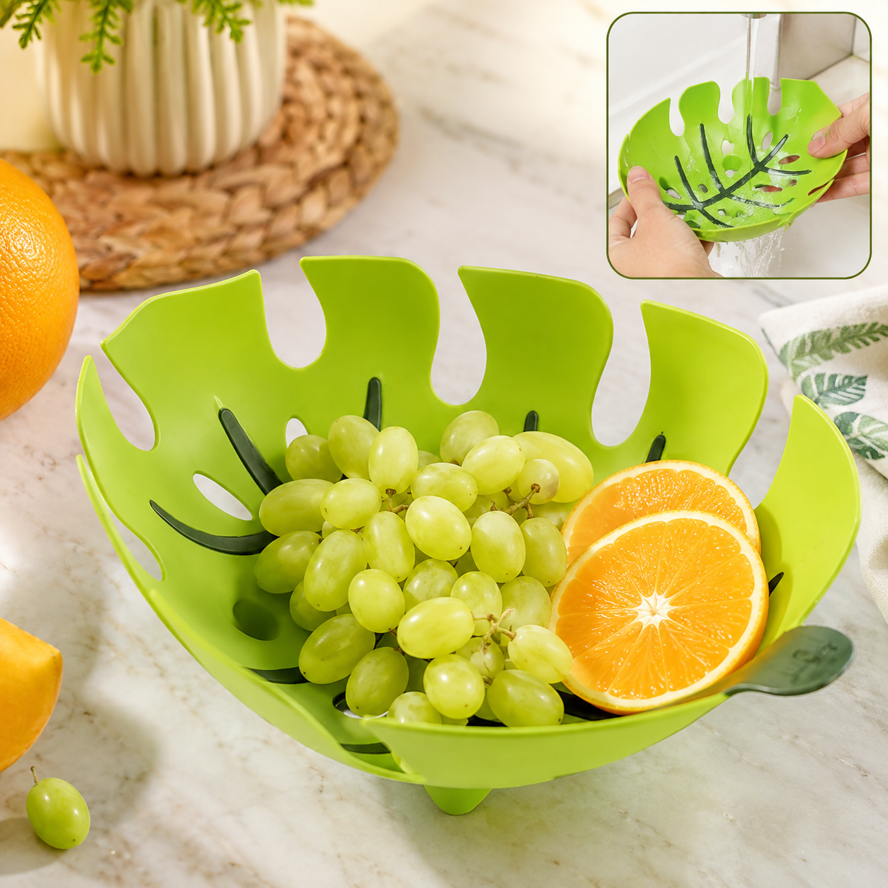 A green leaf-shaped plastic fruit basket filled with green grapes and orange slices on a white marble countertop.