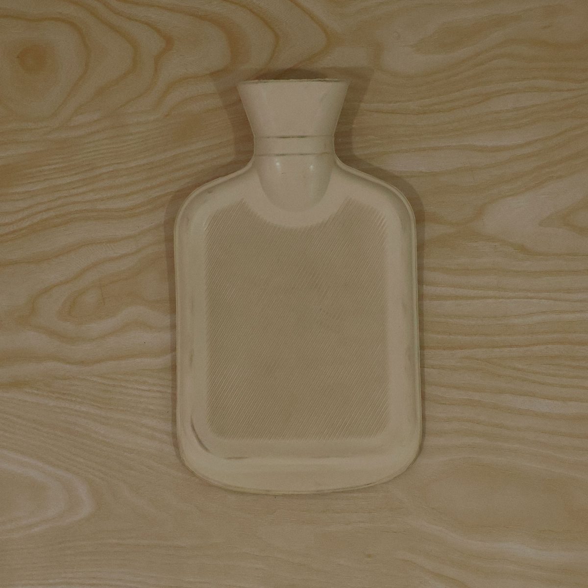 A light yellow hot water bottle with a subtle ribbed pattern lies flat on a light wooden surface.