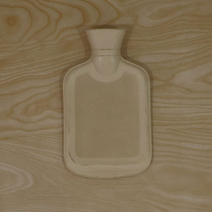 A light yellow hot water bottle with a subtle ribbed pattern lies flat on a light wooden surface.