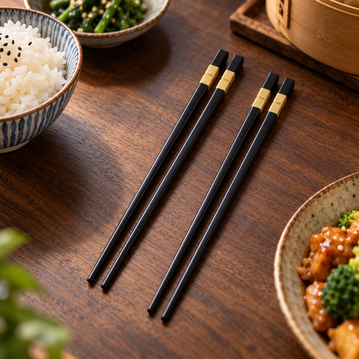 Two pairs of black and gold chopsticks rest on a wooden table next to bowls of rice and other Asian dishes.