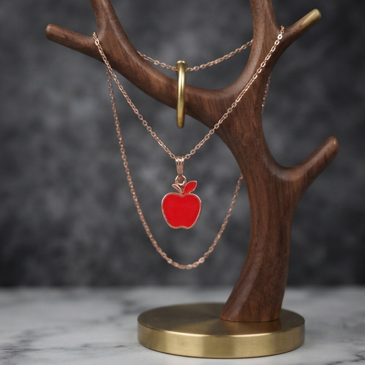 A rose gold necklace with a vibrant red apple pendant hangs on a dark wooden jewelry stand with a brass base.