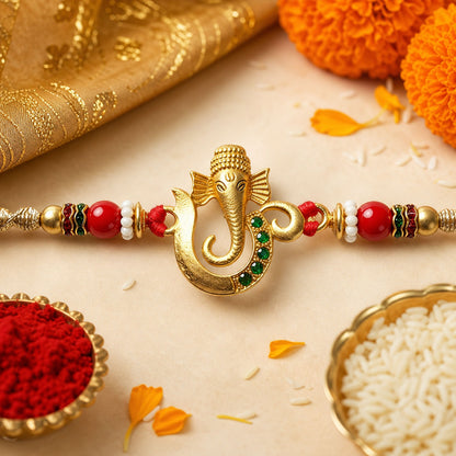 A gold Ganesha charm with green gems is centered on a red and gold beaded thread, surrounded by marigolds, rice, and kumkum