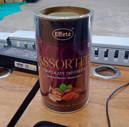 An Effete assorted chocolaty dryfruits can on a light brown wooden desk, next to a black power adapter.