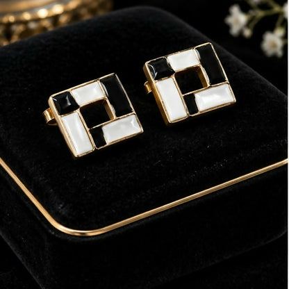 Two black and white square earrings with gold trim on a velvet box.