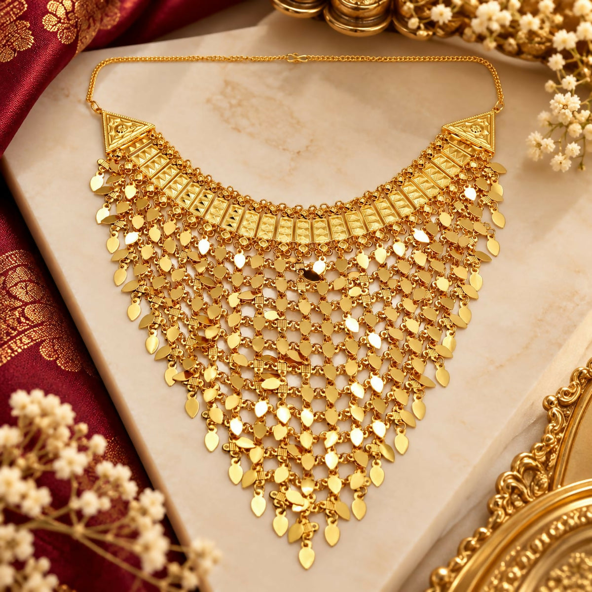 Golden bib necklace with intricate leaf-shaped charms and a detailed upper band, displayed on a white surface with red