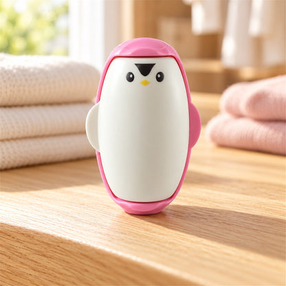 A pink and white penguin-shaped lint roller stands on a wooden surface, with folded towels in the background.