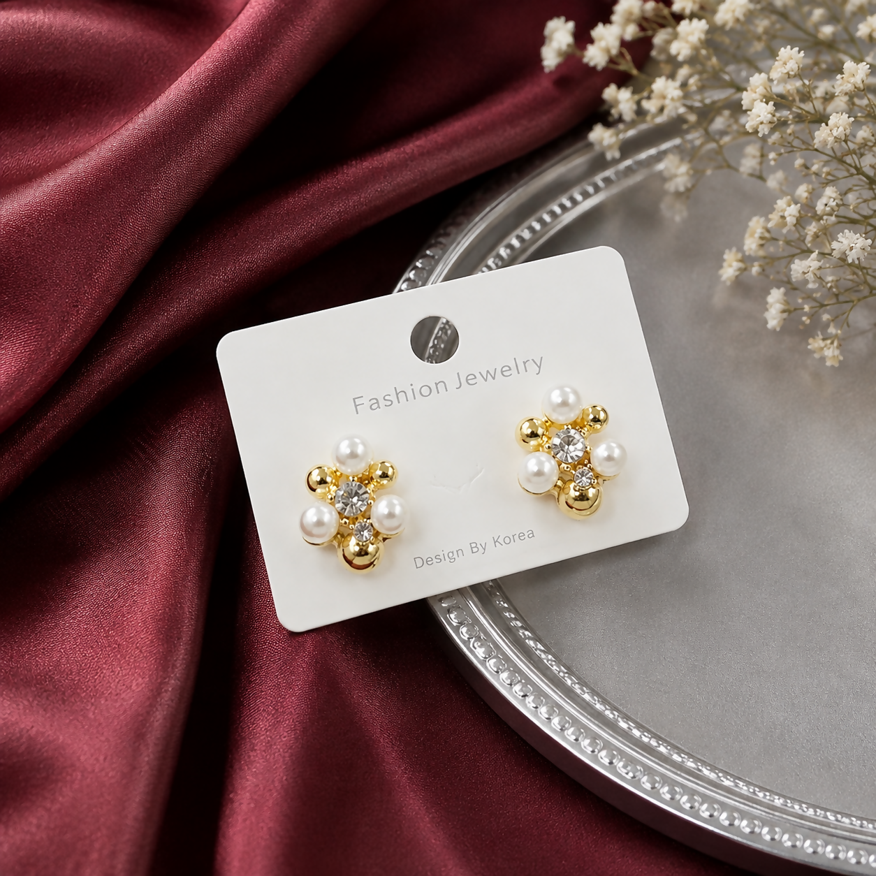 A pair of gold-plated floral stud earrings with crystals and mini pearls on a white card, set against a burgundy satin