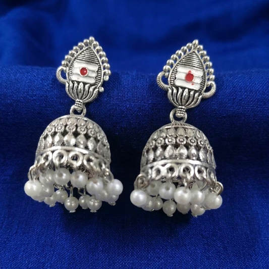 A pair of oxidized silver jhumka earrings with white enamel and red dot, featuring pearl clusters on a blue fabric.