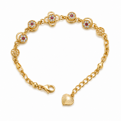 A gold-plated bracelet with floral charms, red and clear gemstones, and a heart-shaped dangle is displayed on a white