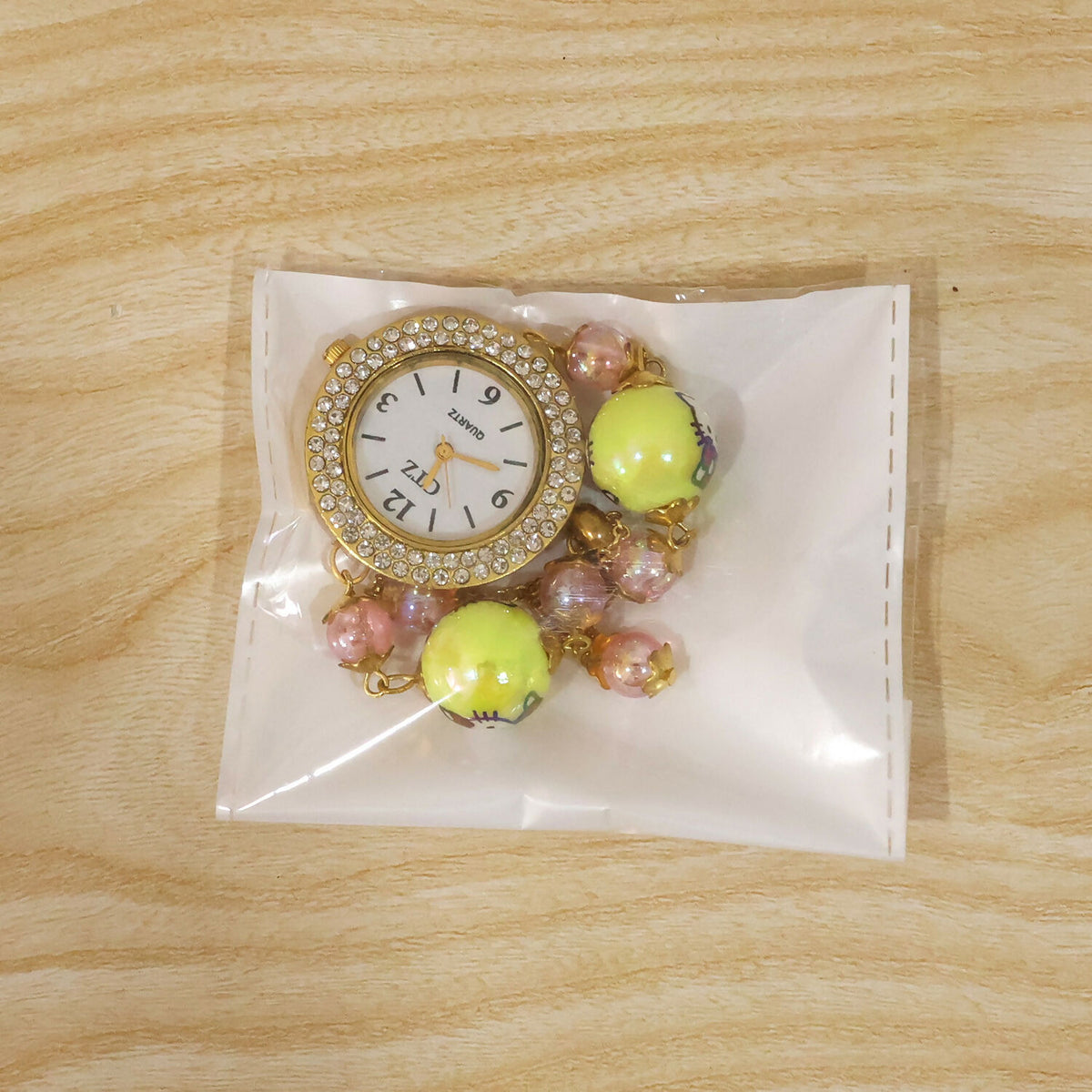 A gold-tone bracelet watch with a rhinestone dial, yellow cartoon character beads, and smaller pink beads, sealed inside a