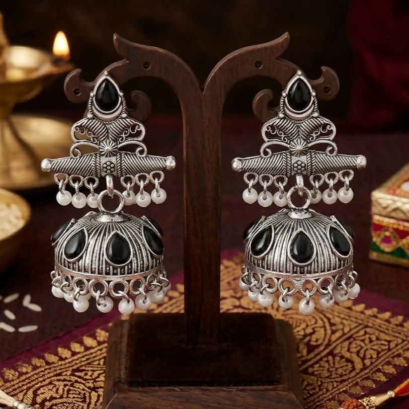 The oxidized silver jhumka earrings with black stones and white pearls are displayed on a dark wooden jewelry stand amidst