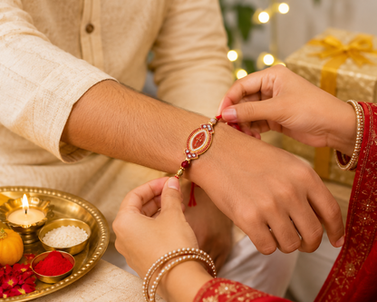 A person in a red garment ties a red and gold bracelet with an oval centerpiece onto another person's wrist, with a gift box