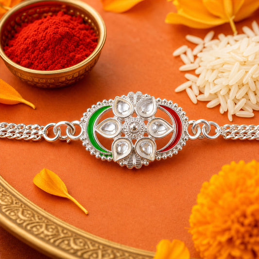 A silver rakhi with a central flower design featuring clear stones and green and red enamel accents, surrounded by rice and