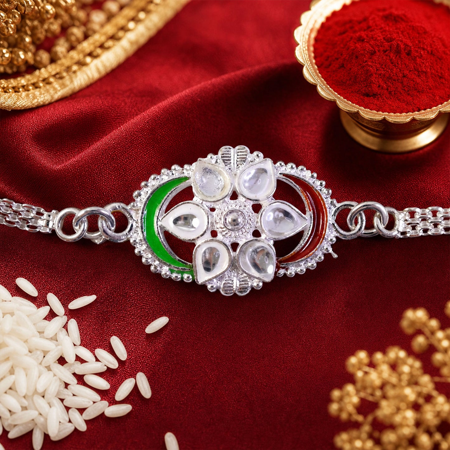 A silver rakhi with a flower design, clear stones, and green and red enamel, resting on a red satin cloth with rice and gold