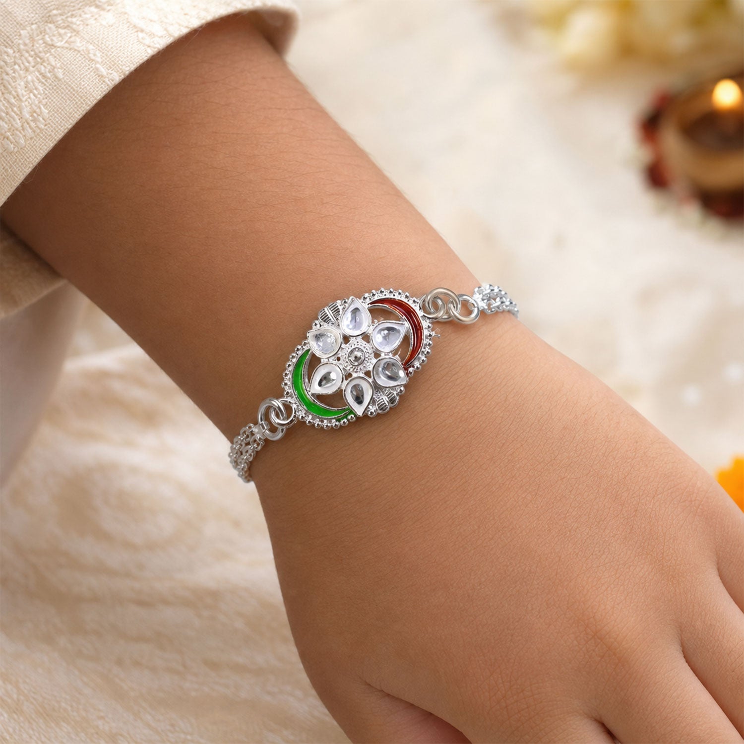 A silver rakhi with a tricolor chain link design and clear stones on a person's wrist, with a blurred background.