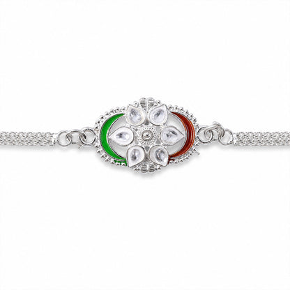 A silver rakhi with a central flower motif adorned with clear stones and green and red enamel, presented on a plain white