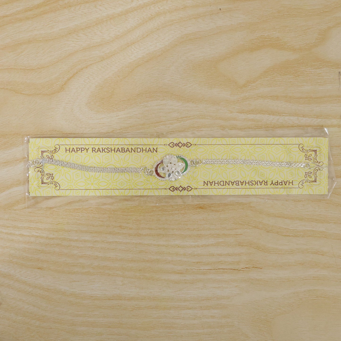 A silver rakhi with a tricolor chain link design and clear stones, packaged on a yellow card with