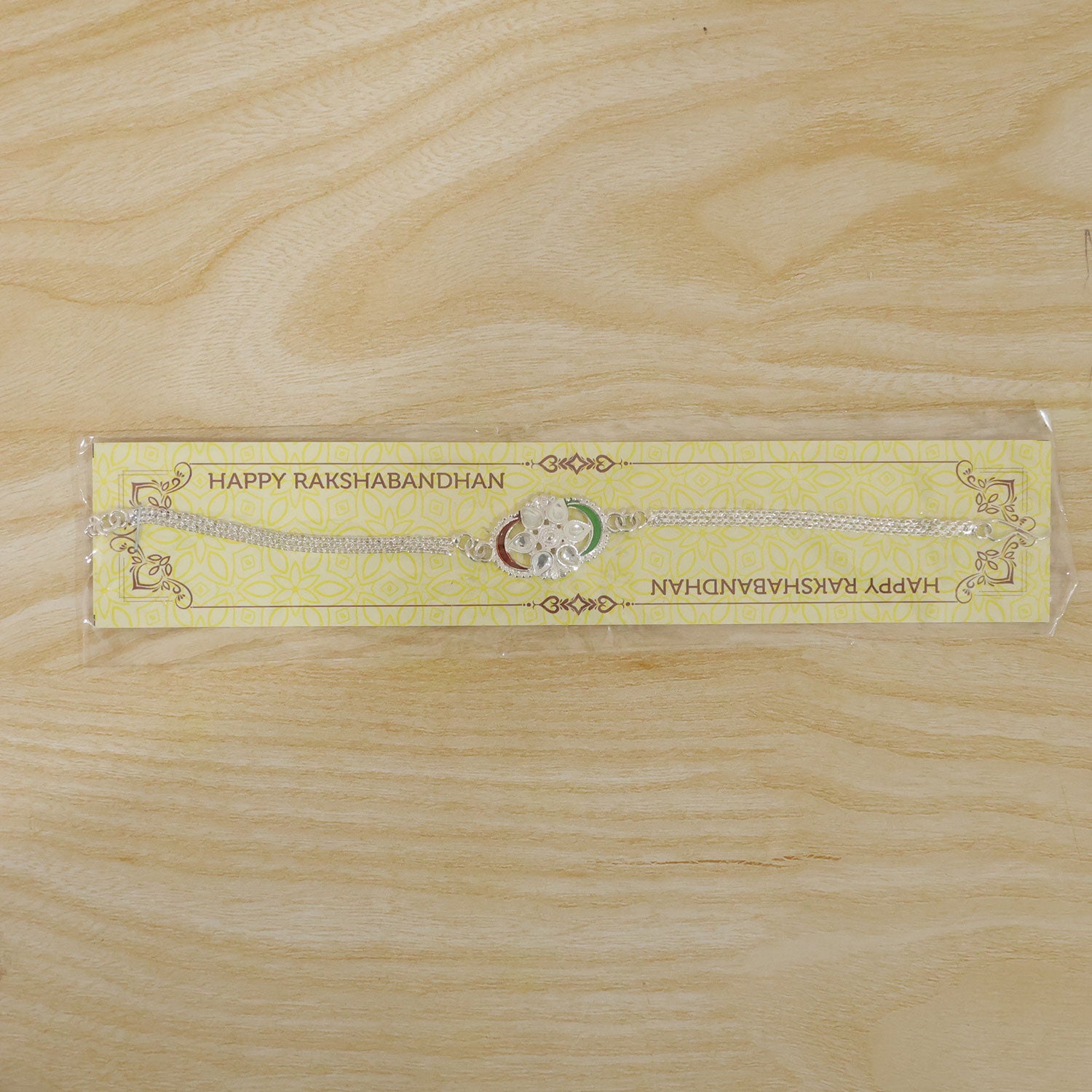 A silver rakhi with a tricolor chain link design and clear stones, packaged on a yellow card with