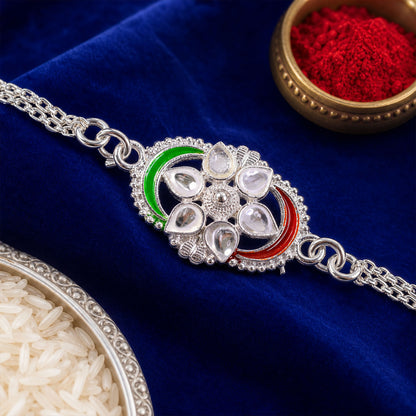 A silver rakhi with clear stones and green and red enamel on a blue velvet surface, with a bowl of red powder and rice.