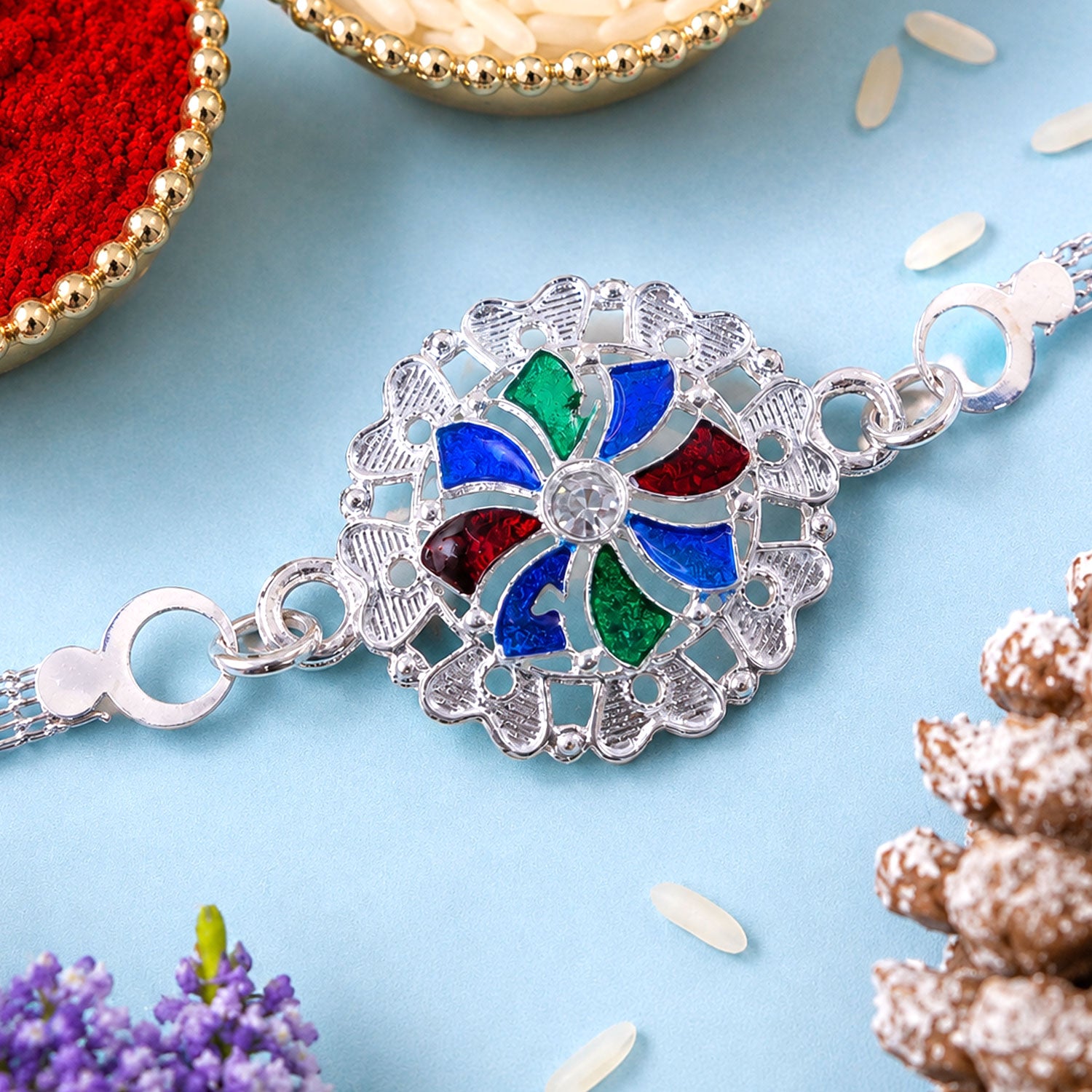 A silver chain bracelet with a round, colorful enamel and crystal centerpiece is shown on a light blue surface with