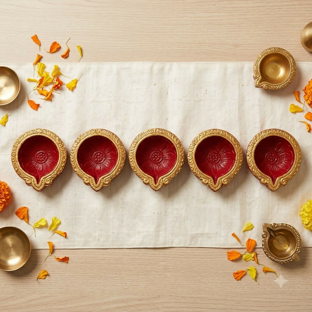 Overhead view of five red and gold diyas on a white fabric runner, flanked by brass bowls and orange petals.