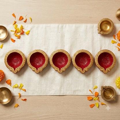 Overhead view of five red and gold diyas on a white fabric runner, flanked by brass bowls and orange petals.