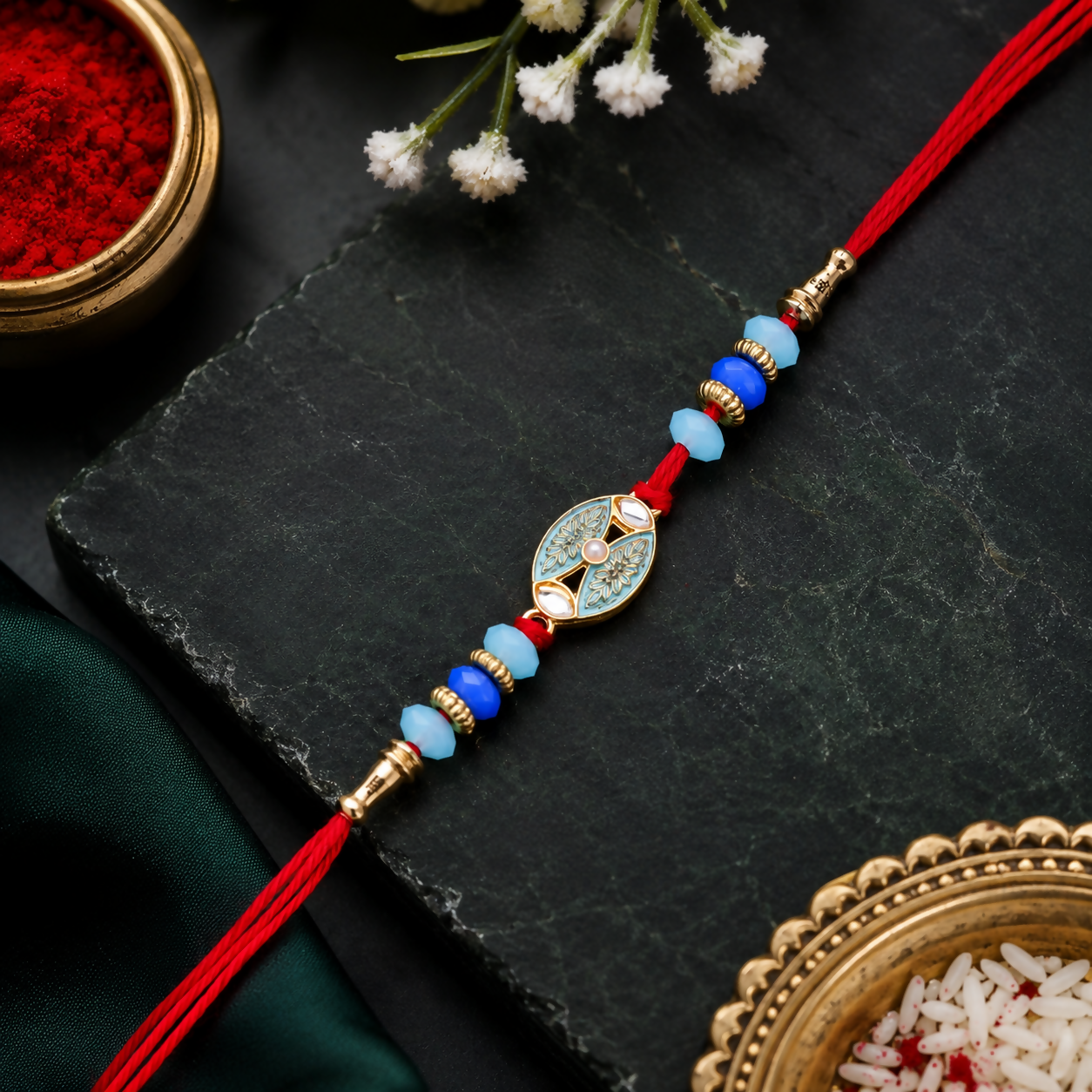 A red thread bracelet with blue and light blue beads and a central golden charm with a light blue design, lies on a dark
