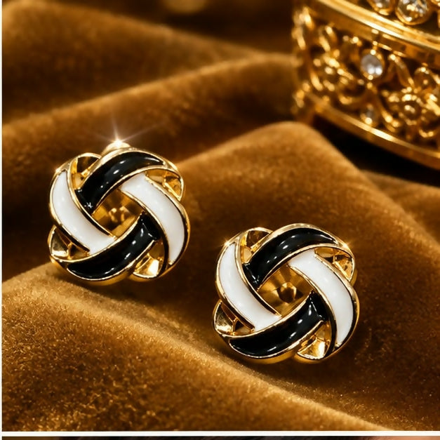 A pair of black and white enamel knot stud earrings rest on a textured brown fabric.