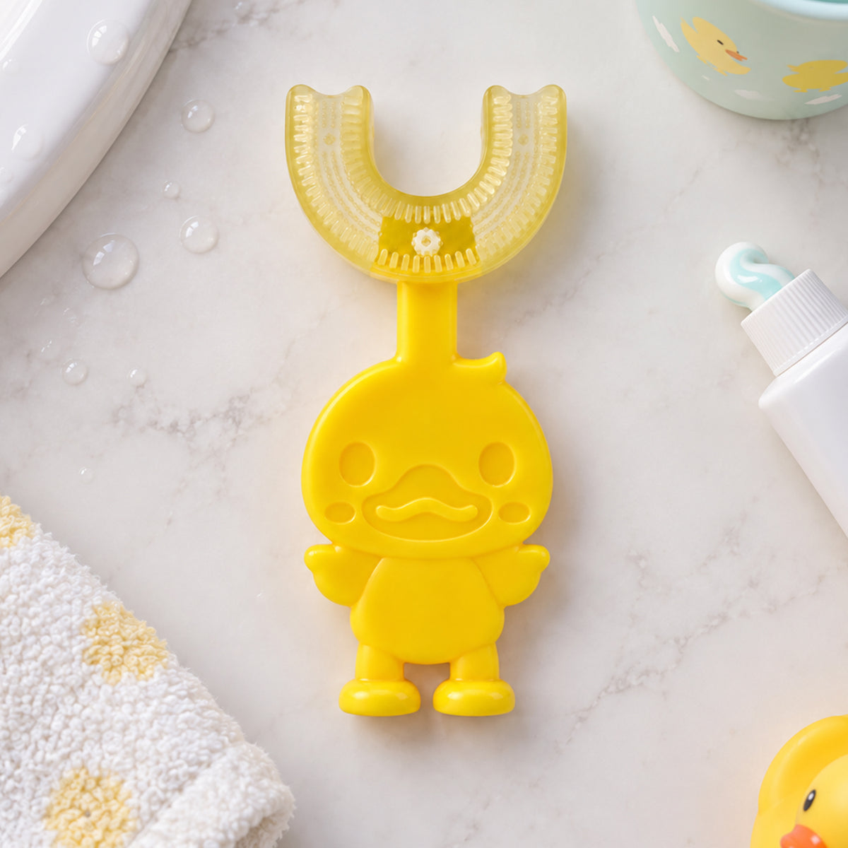 A yellow duck-shaped U-shaped toothbrush lies on a white marble counter with a towel and toothpaste.