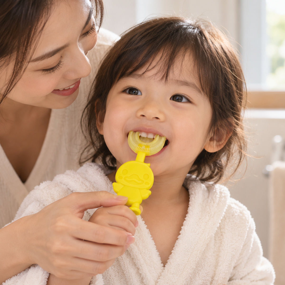 A smiling adult helps a young child brush their teeth with a yellow duck-shaped U-shaped toothbrush.