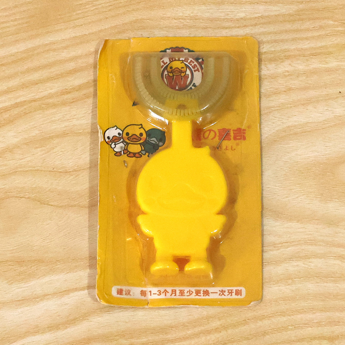 A yellow duck-shaped U-shaped toothbrush in worn clear plastic packaging with Asian text on a wooden surface.