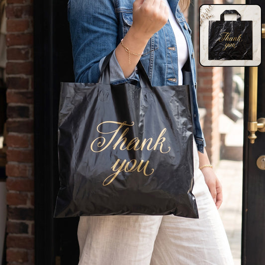 A person in a denim jacket holds a black plastic 'Thank You' bag with gold script, outdoors.