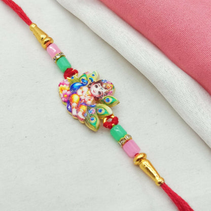 The Rakhi, featuring a colorful Krishna and peacock feather charm, is displayed on a white surface with a pink fabric fold.