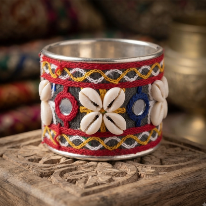 A wide silver cuff bracelet with colorful embroidered patterns, small round mirrors, and white cowrie shells sits on a
