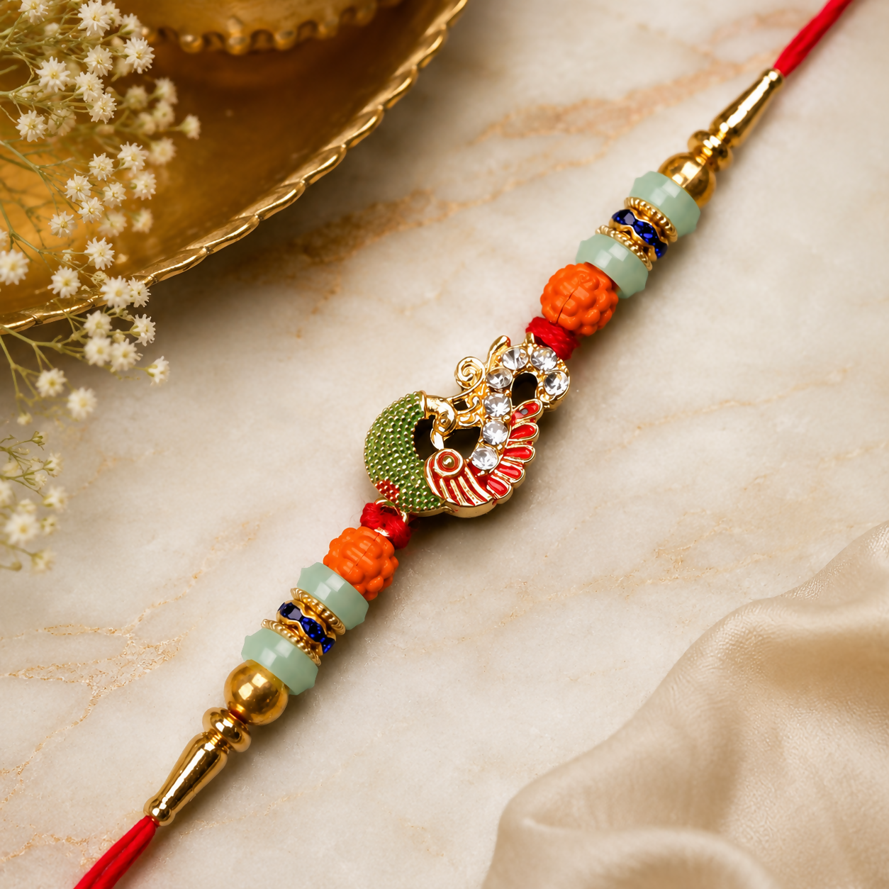 A red thread with a gold and green peacock charm, orange and mint green beads, and gold accents, rests on a white marble