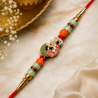 A red thread with a gold and green peacock charm, orange and mint green beads, and gold accents, rests on a white marble