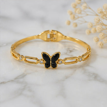 A gold-tone bracelet with a black butterfly charm and crystal accents rests on a white marble surface.