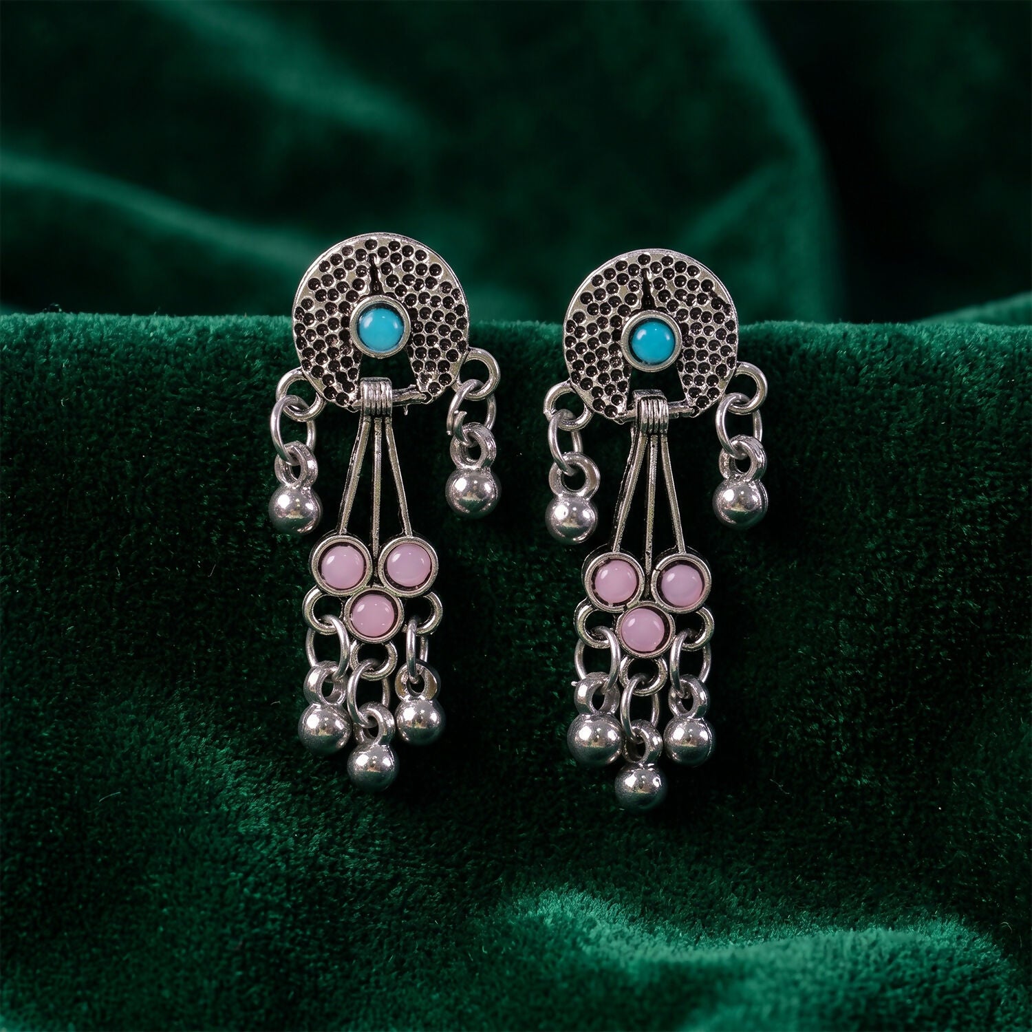 The oxidised silver long drop earrings with pink and turquoise stones and ghungroo bells are displayed on a dark green