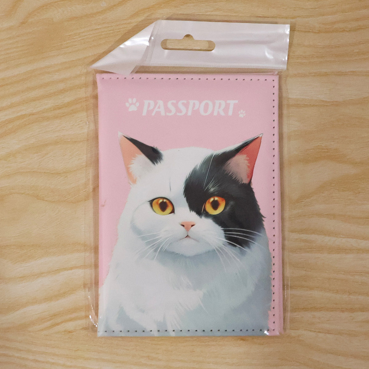The pink passport holder with a cat design is presented in clear plastic packaging against a light wooden background.