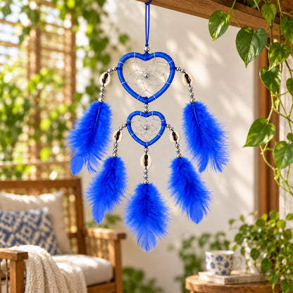 A blue heart-shaped dream catcher with feathers hangs outdoors on a wooden beam, surrounded by green foliage.