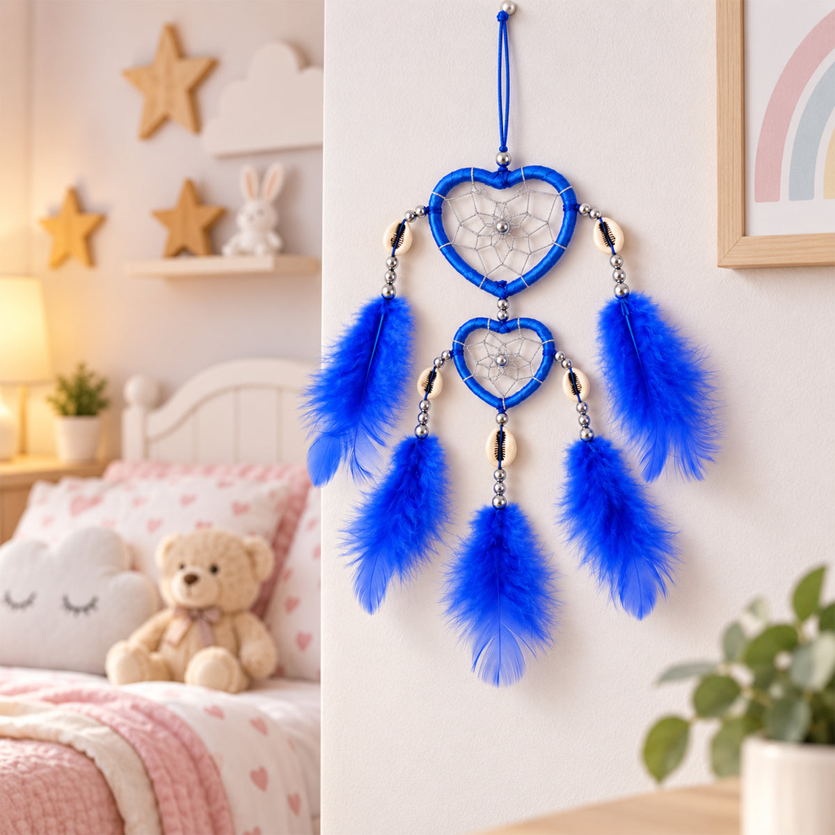 A blue heart-shaped dream catcher with feathers hangs on a white wall in a child's bedroom with a teddy bear and bed visible.