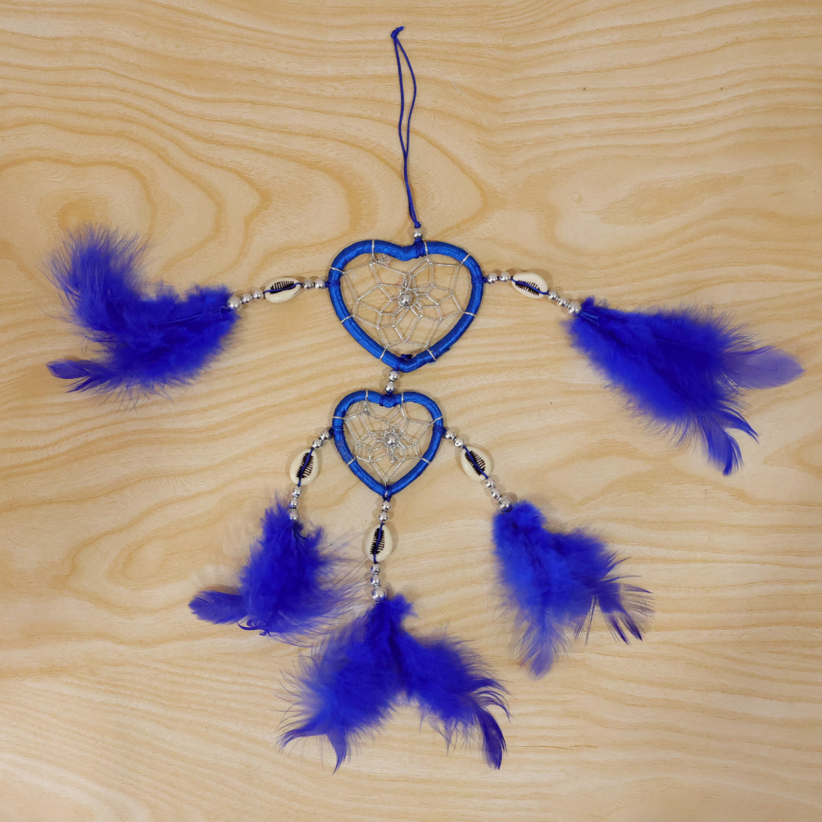 A blue heart-shaped dream catcher with feathers and silver accents lies flat on a light wooden surface.