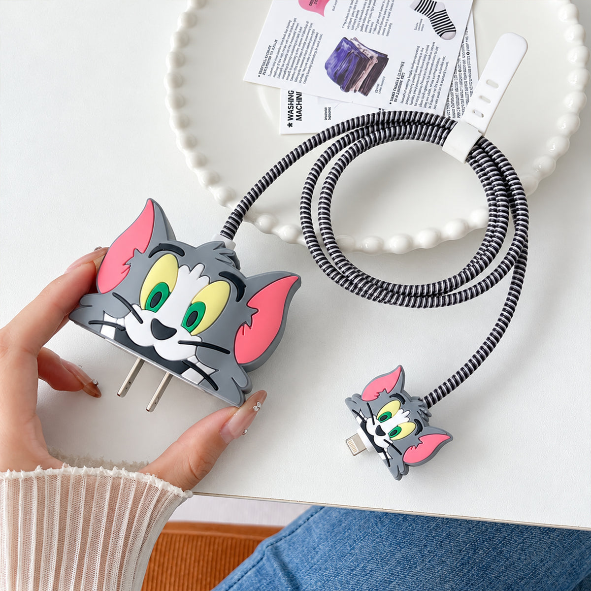 A hand holds a gray Tom cat silicone charger cover with a matching cable protector attached to a coiled cable.