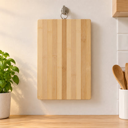 A rectangular bamboo chopping board hangs vertically on a silver hook against a white kitchen wall.